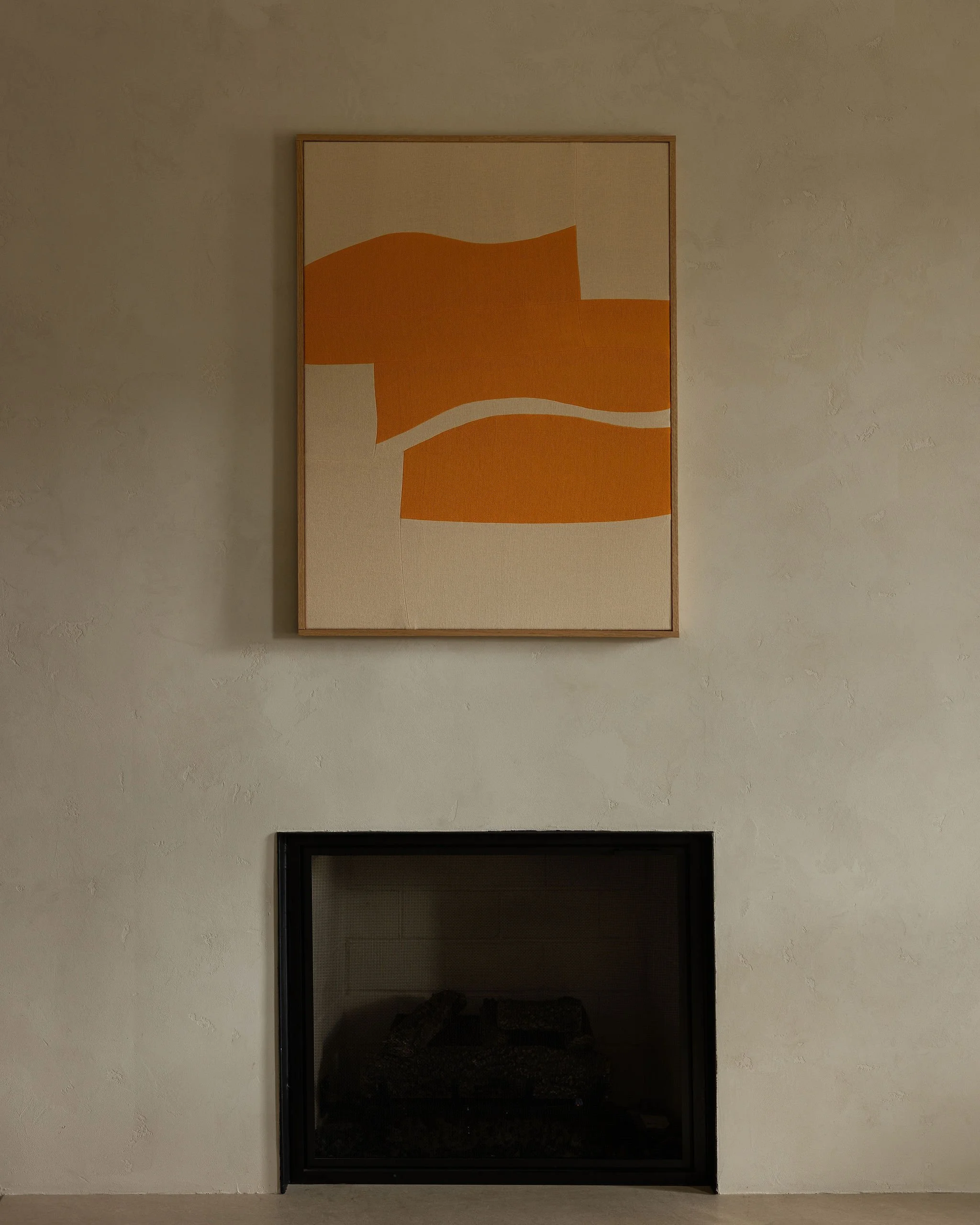 A softly plastered fireplace wall in the front living room, with a framed abstract work in warm orange and cream hung above a minimal black firebox.