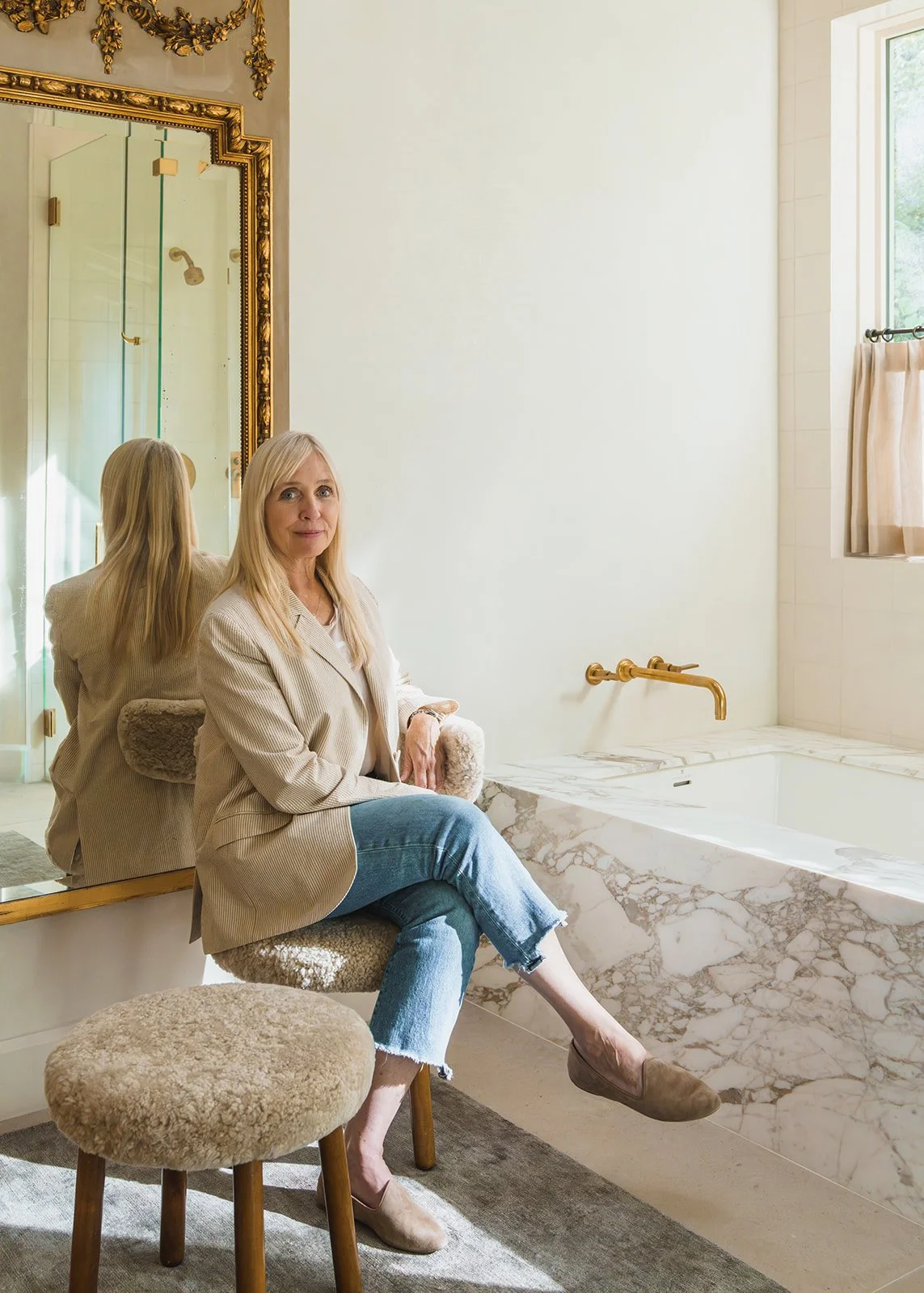Laura Roberts seated on a stool beside a marble tub, reflected in a gilt-framed mirror.