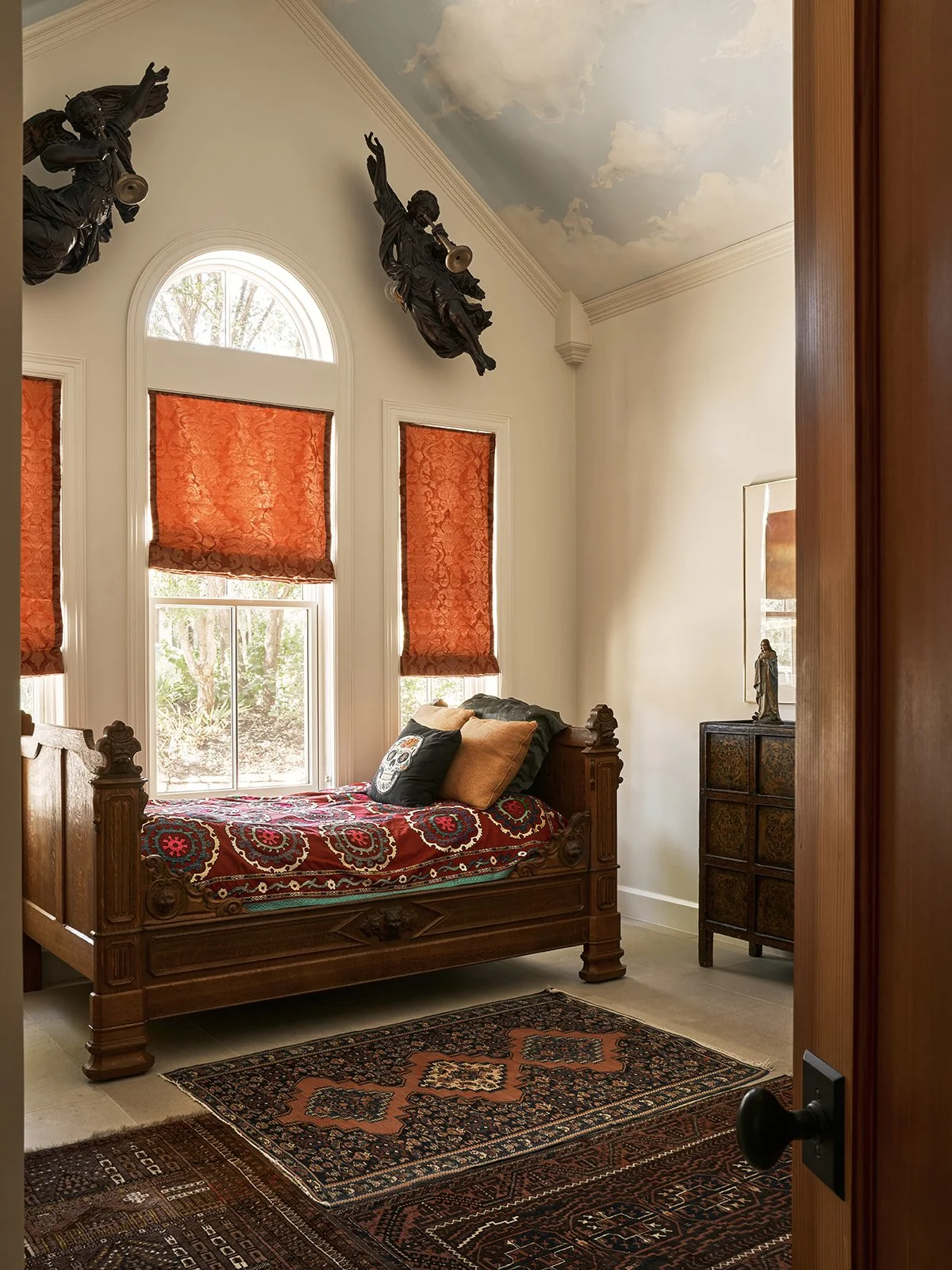 “Cloud room” guest bedroom featuring hand-painted cloud ceiling murals, a vintage wood daybed, layered antique rugs, silk brocade window shades, carved angel sculptures, and a Tibetan chest.