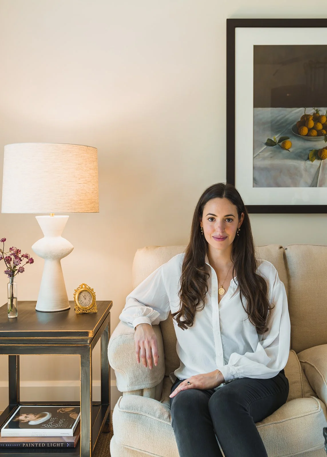 Kristen Thorsen seated on a sofa beside a side table with a sculptural lamp and a still-life painting above.