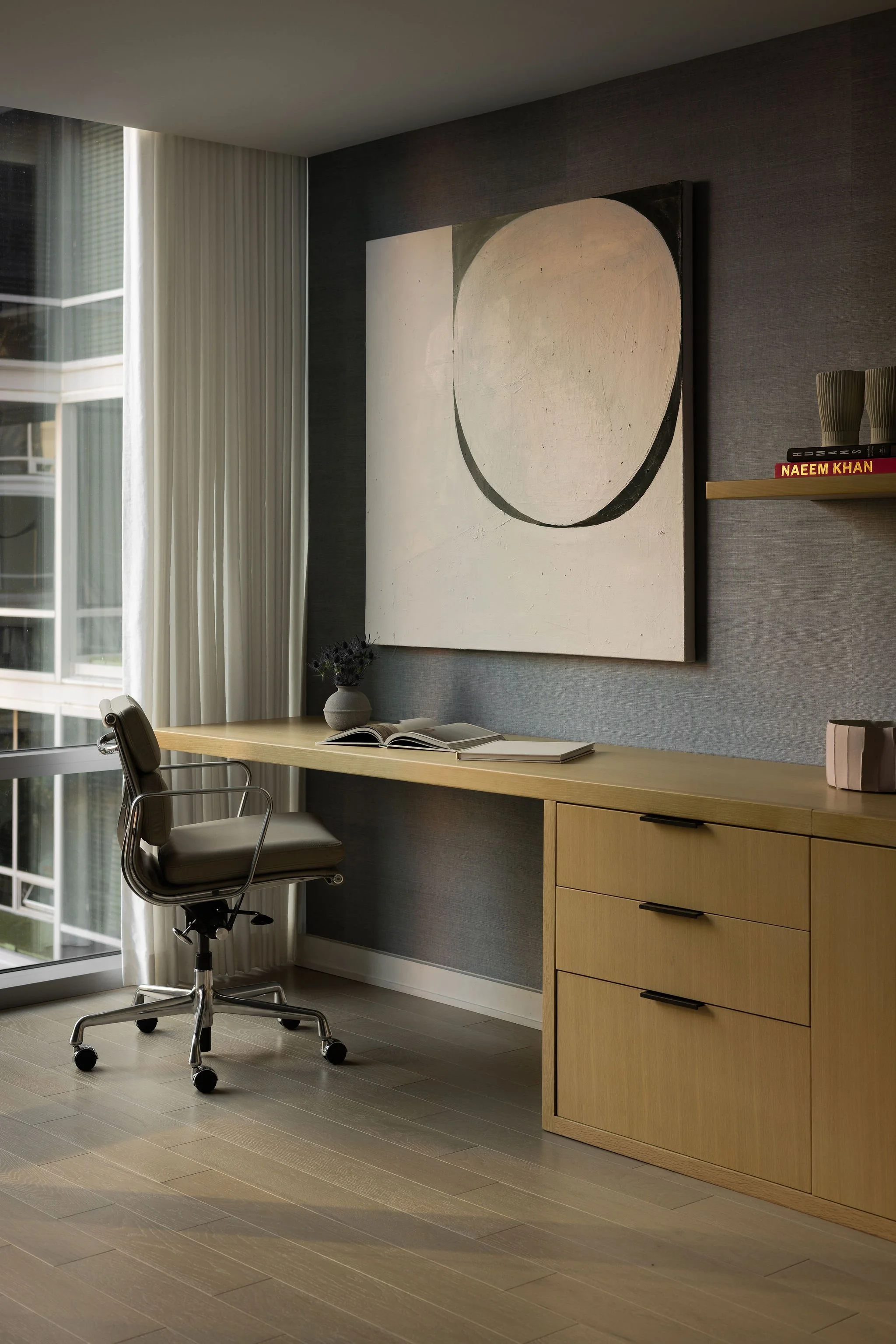 Home office in a Georgetown, DC condo with a floating oak desk and drawers, a black leather task chair, and a large abstract painting of a pale ovoid on a charcoal grasscloth wall beside a curtained window.