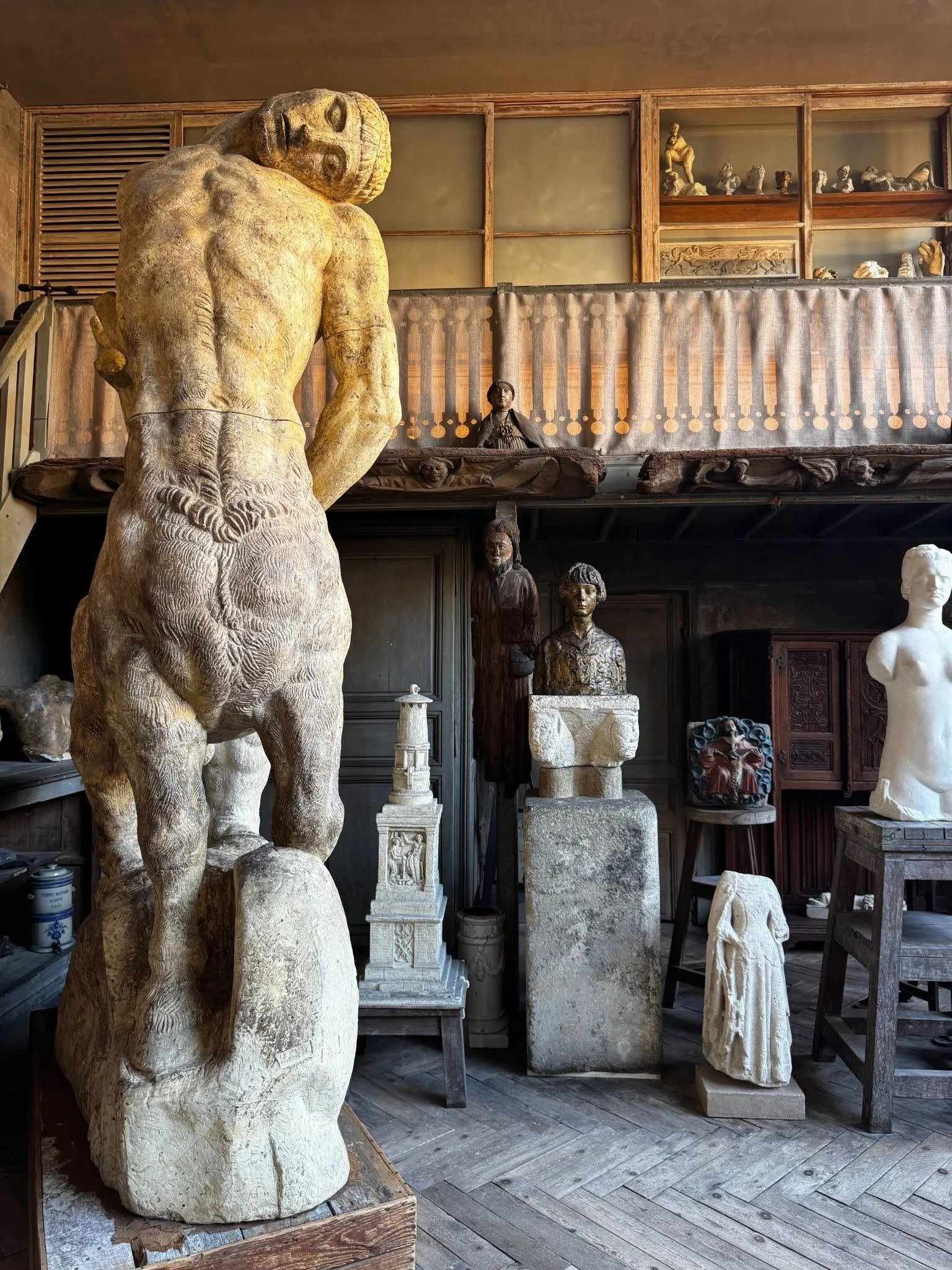 A sculptor's atelier with a large ochre standing figure seen from behind, surrounded by carved busts and fragments on stone plinths and a herringbone wood floor.
