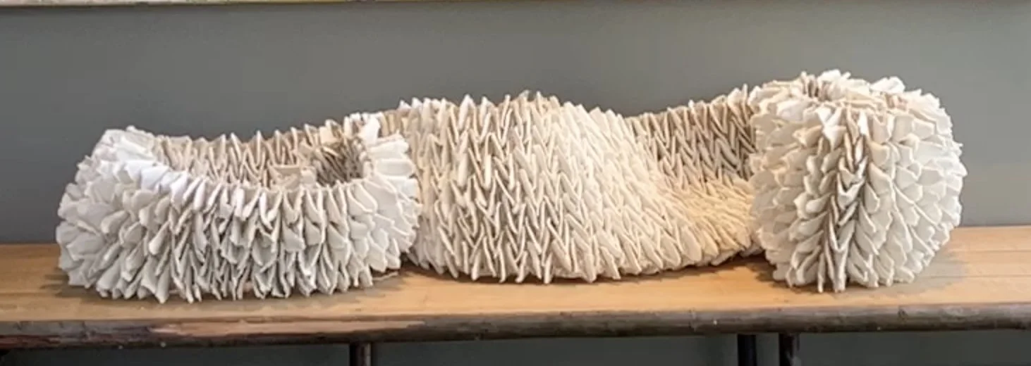 A long undulating sculpture of ruffled white porcelain petals threaded on cord, resting on a wooden bench.