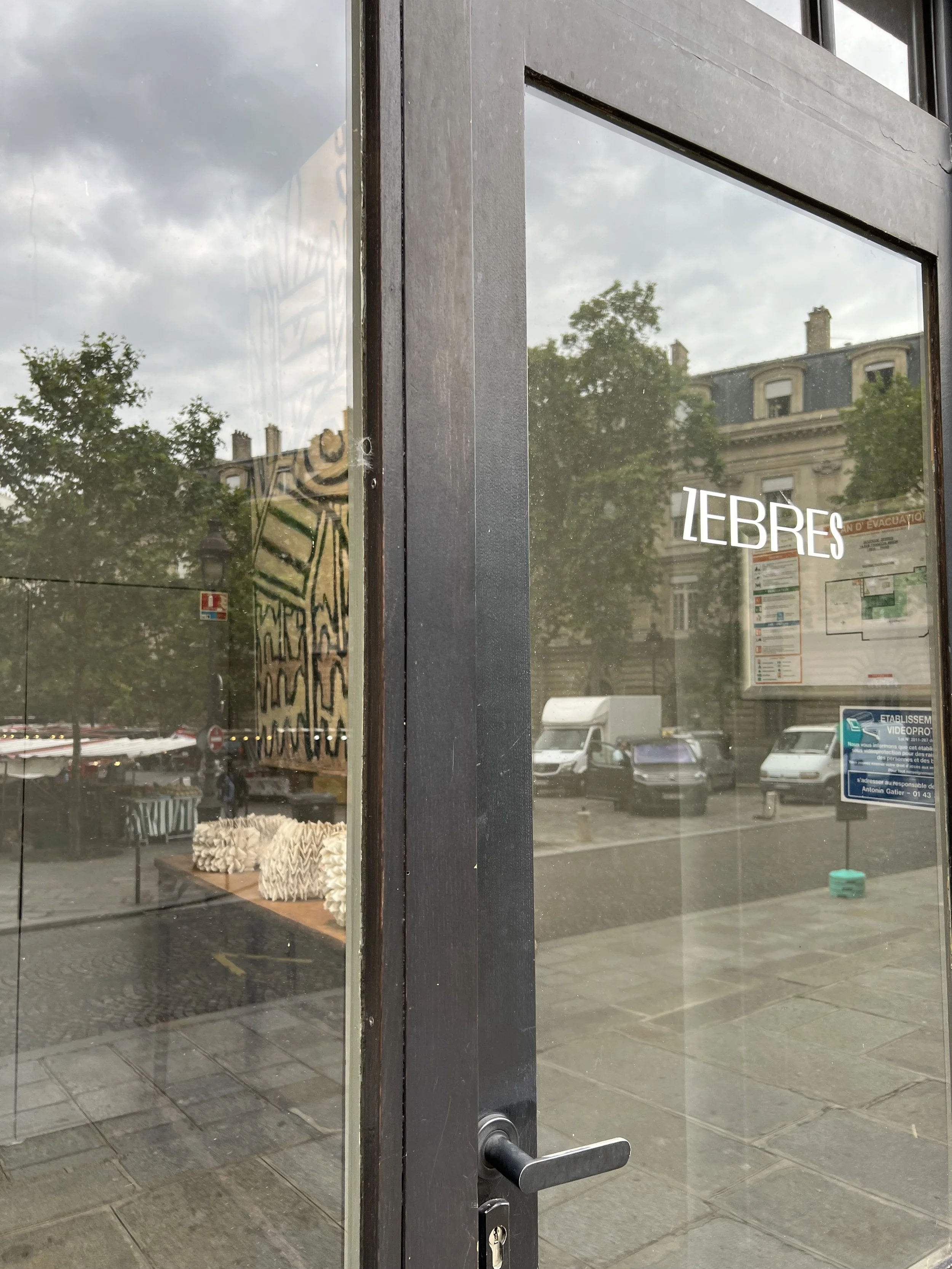 Glass entrance door of the ZEBRES gallery in Paris, reflecting a tree-lined street and market stalls, with a pale sculptural work visible inside.