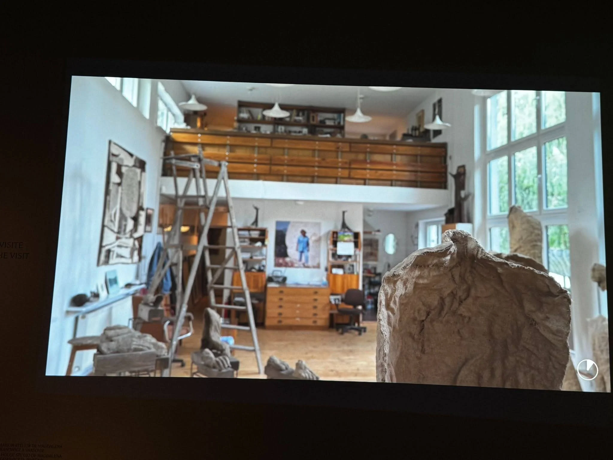 A projected photograph of a sculptor's house-studio — a tall light-filled room with a library ladder, mezzanine storage, and works in progress — displayed behind a rough stone sculpture.