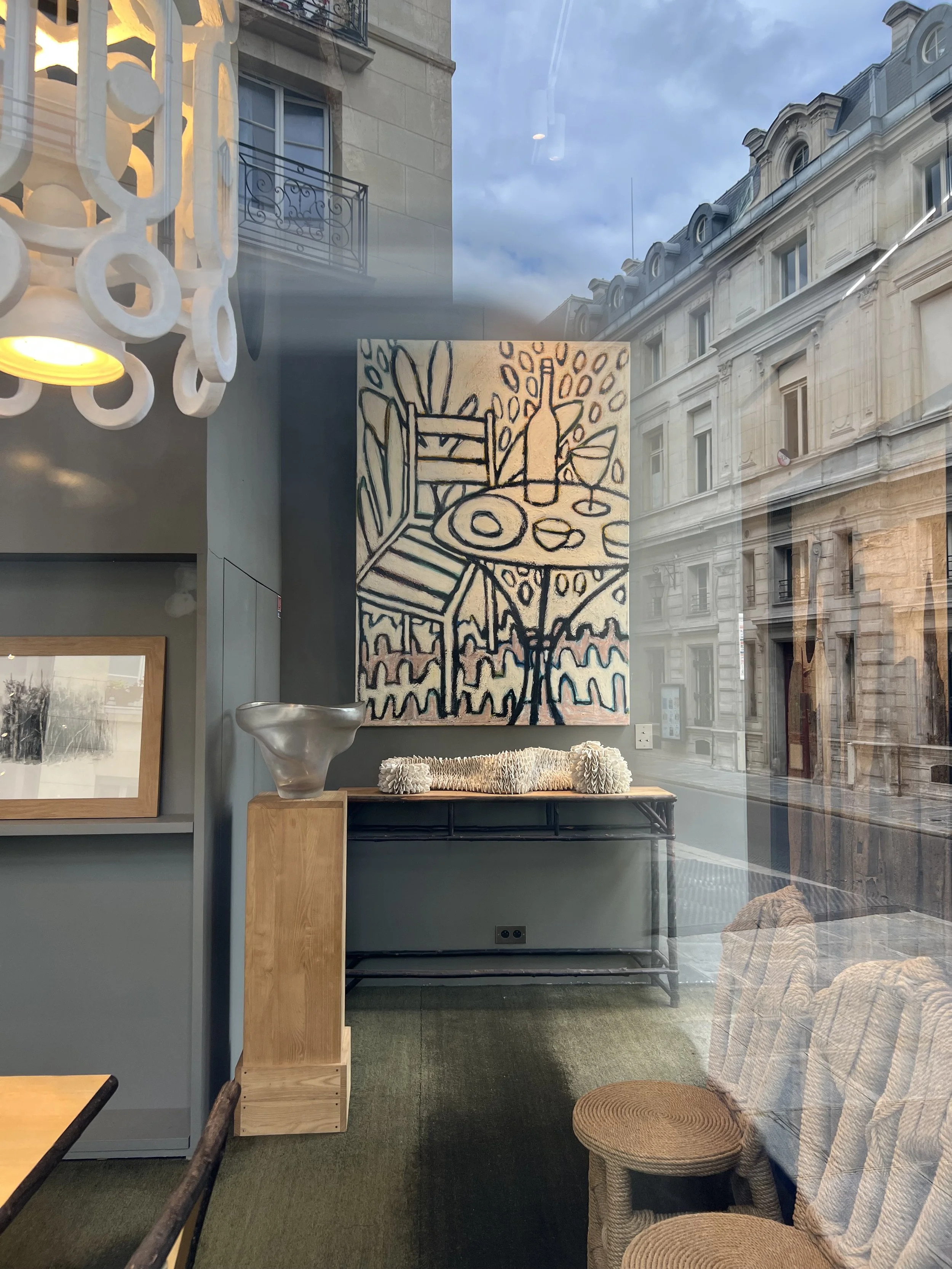 A gallery window with an abstract still-life painting above a console holding a silver vessel and a white pleated ceramic sculpture, Parisian buildings reflected in the glass.