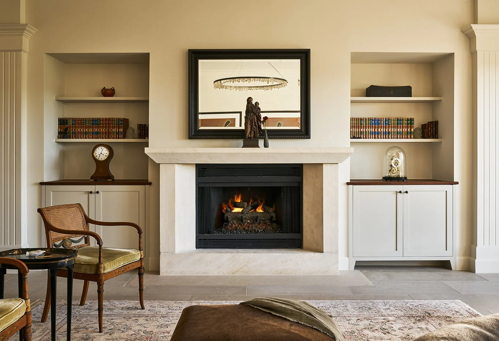 Cream-toned living room with a limestone fireplace and lit gas fire, a black-framed mirror above the mantel reflecting a crystal ring chandelier, niches of leather-bound books, and antique caned armchairs on a patterned rug.